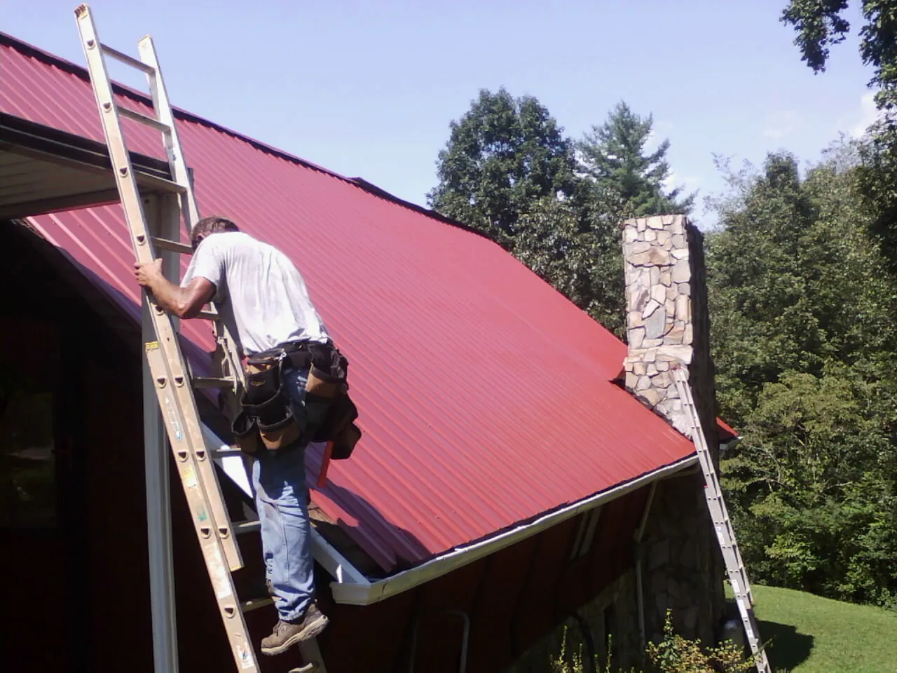 Roofer on a ladder beside a red metal roof and stone chimney — Soffit & Fascia Repair by Roofing Brothers in Old Greenwich